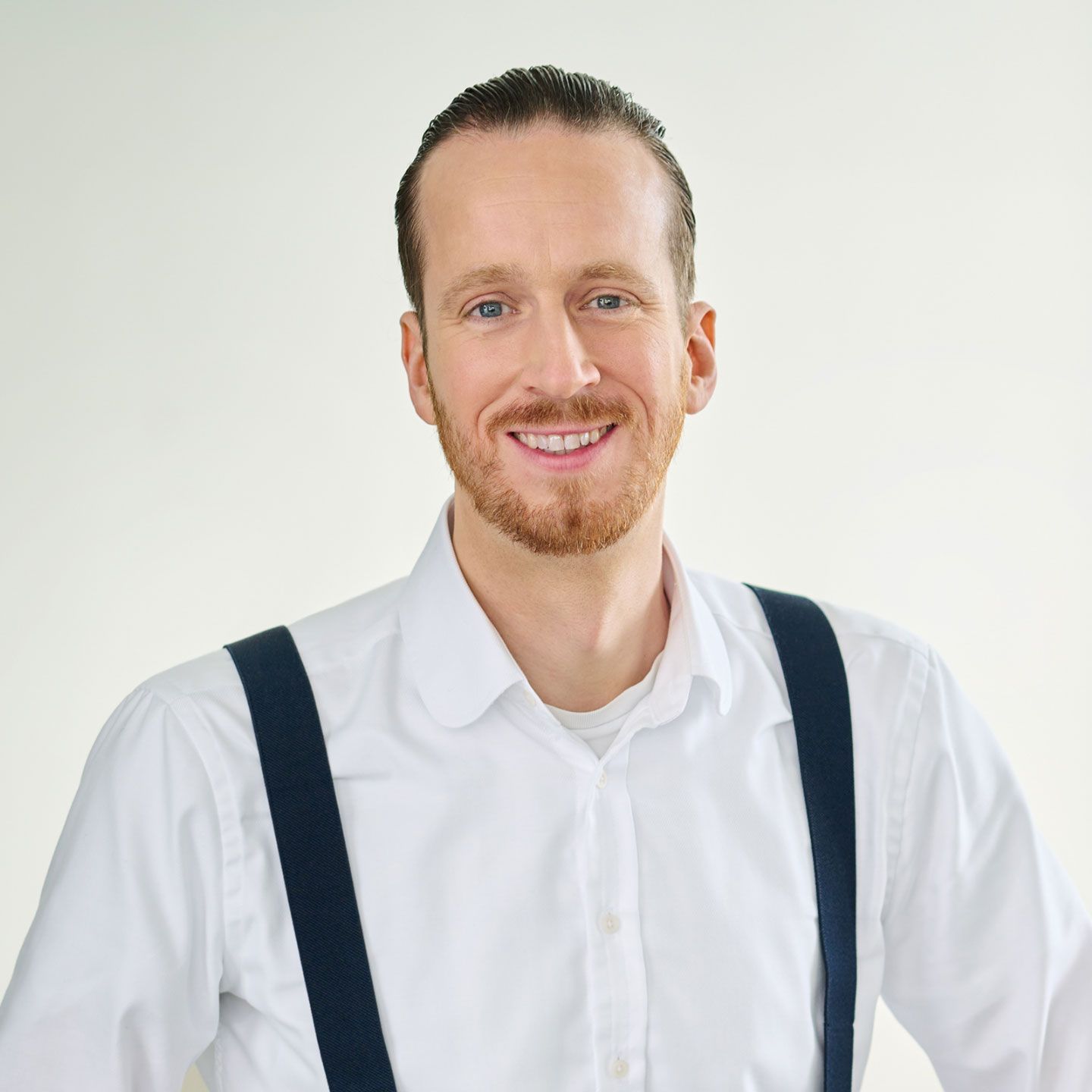 Sebastian Pollmer, mężczyzna stoi w białej koszuli z niebieskimi szelkami w jasnym pomieszczeniu. 