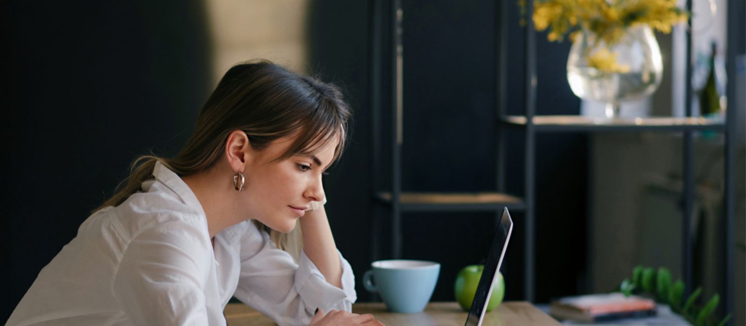 Młoda kobieta skupiona pracuje na laptopie przy biurku, obok leżą notatnik i filiżanka.