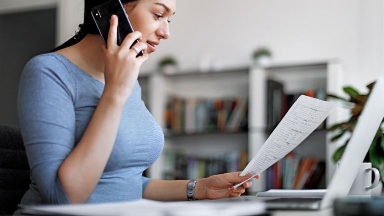 Kobieta w niebieskiej bluzce rozmawia przez telefon komórkowy, trzymając fakturę w rękach.