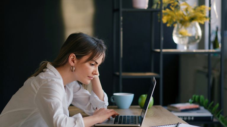 Młoda kobieta skupiona pracuje na laptopie przy biurku, obok leżą notatnik i filiżanka.
