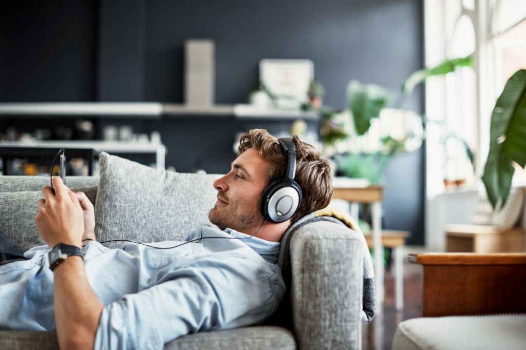 Młody mężczyzna leży na kanapie w swoim salonie, ma na uszach słuchawki i z uśmiechem patrzy na smartfona, słuchając muzyki lub podcastu.