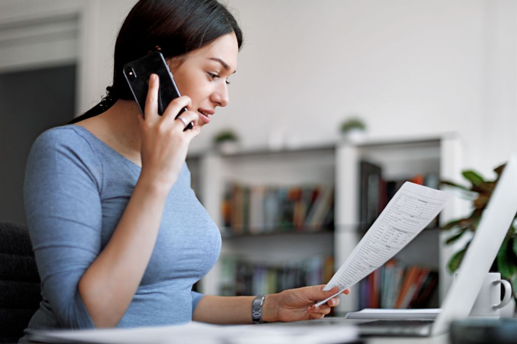 Kobieta w niebieskiej bluzce rozmawia przez telefon komórkowy, trzymając fakturę w rękach.