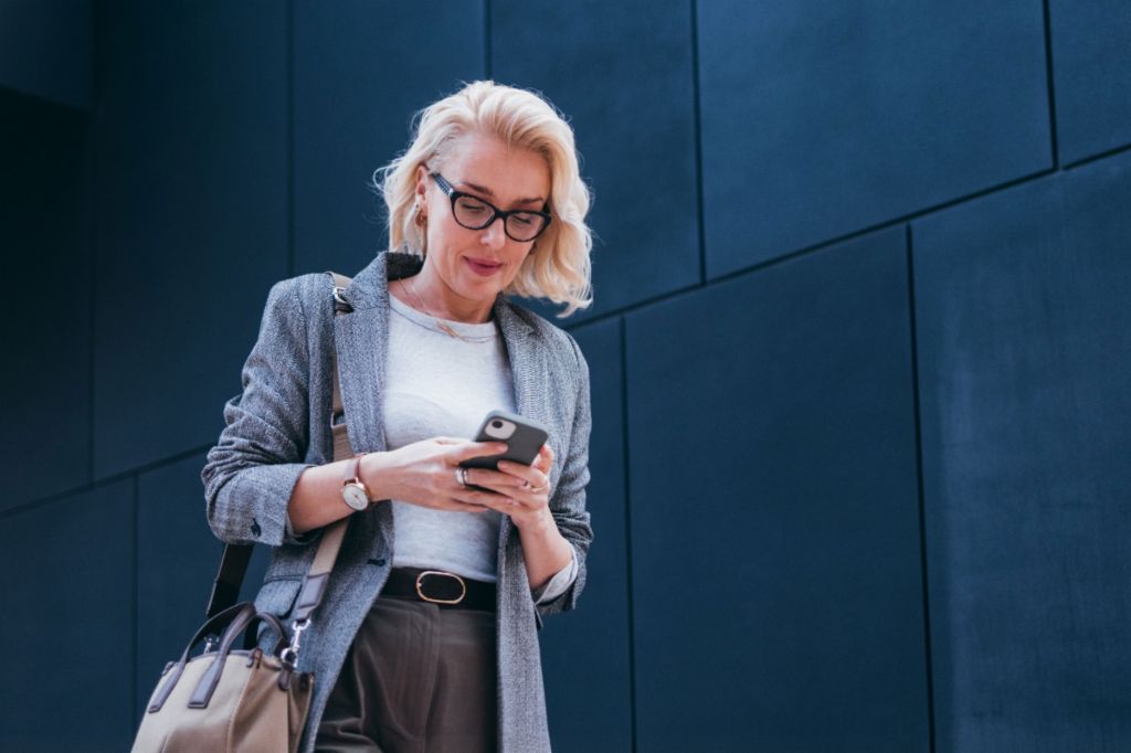 Blondynka w szarej marynarce stoi przed nowoczesnym budynkiem i uśmiecha się, patrząc na swój smartfon.