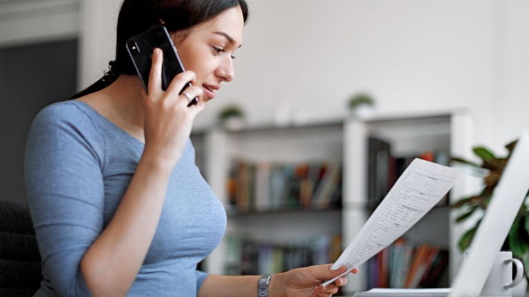 Kobieta w niebieskiej bluzce rozmawia przez telefon komórkowy, trzymając fakturę w rękach.