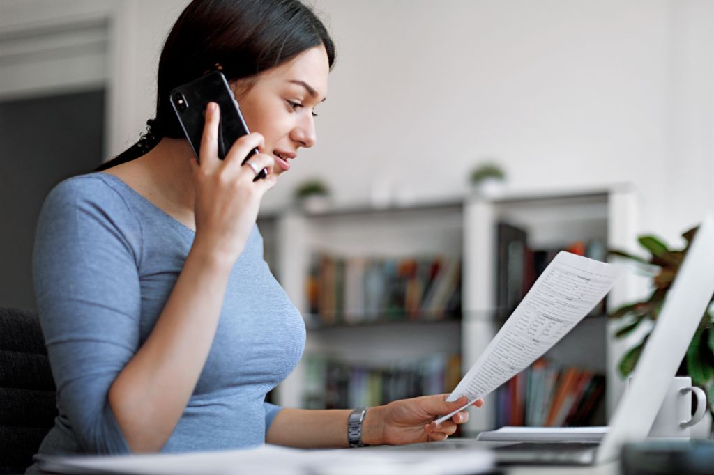 Kobieta w niebieskiej bluzce rozmawia przez telefon komórkowy, trzymając fakturę w rękach.