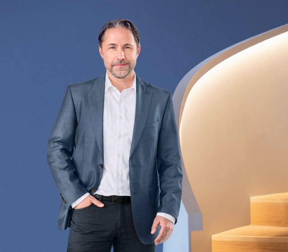 Marwin Ramcke, Dyrektor Generalny Grupy EOS, stoi przed niebieskim tłem, ubrany w ciemną marynarkę. Patrzy bezpośrednio w obiektyw kamery.
