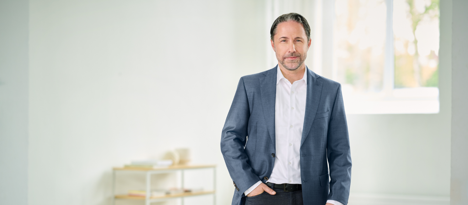 Marwin Ramcke, CEO, stoi w niebieskim garniturze z prawą ręką w kieszeni spodni w jasnym pomieszczeniu. 