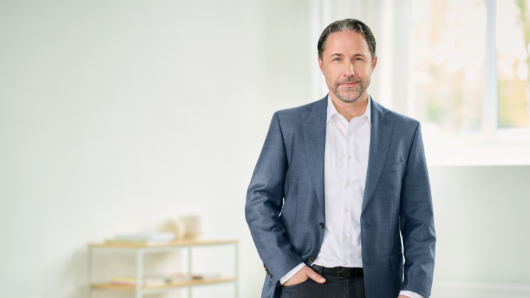 Marwin Ramcke, CEO, stoi w niebieskim garniturze z prawą ręką w kieszeni spodni w jasnym pomieszczeniu. 