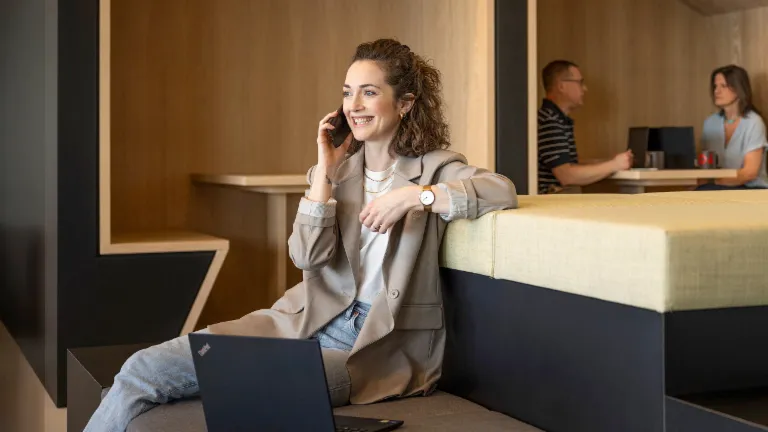 Uśmiechnięta osoba siedzi w nowoczesnej strefie lounge biura, rozmawia przez telefon i pracuje na laptopie. W tle pracują dwie inne osoby.
