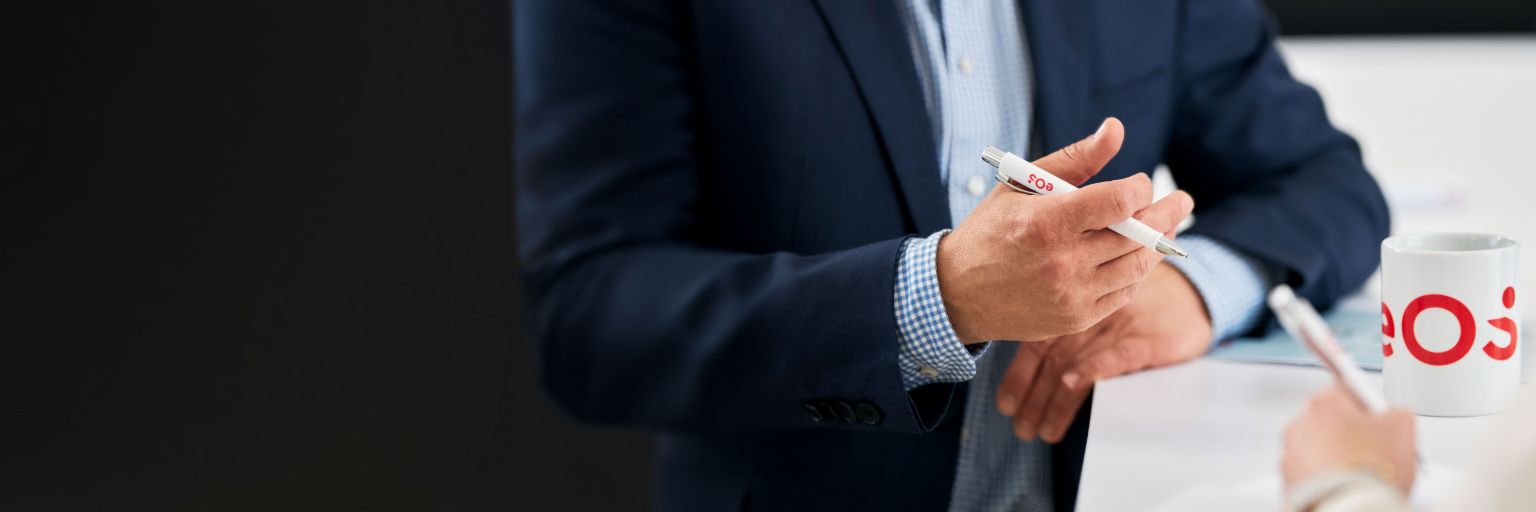 Biznesmen trzymający długopis EOS podczas rozmowy o międzynarodowym zarządzaniu należnościami, kubek EOS na stole - Ekspertyza w windykacji, NPL i rozwiązaniach cyfrowych