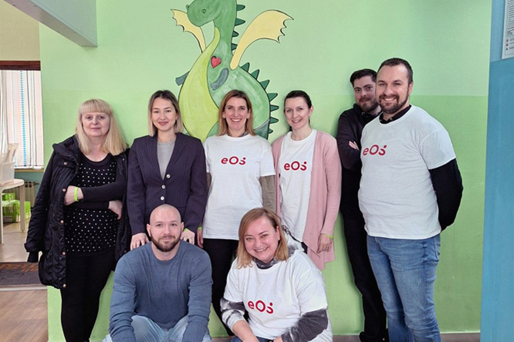 Osiem osób stoi na tle ściany, na której namalowano zielonego smoka. Są ustawione w dwóch rzędach: dwie osoby kucają z przodu, a sześć osób stoi z tyłu. Cztery osoby mają na sobie białe koszulki z czerwonym logo EOS. Grupa uśmiecha się do aparatu. Na podłodze, z przodu po prawej stronie, widać kilka butelek i kartonów z napojami.